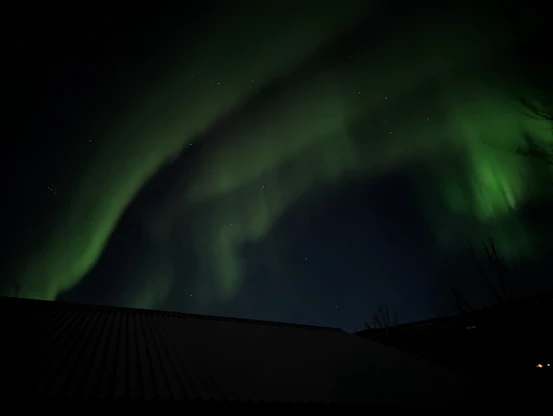 Multiple green Aurora "curtains" in the sky above a wood cabin roof.