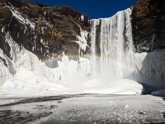 A large waterfall that flows off an almost barren cliff under a deep blue sky. Snow and ice cover the lower parts of the cliffs, and a large rainbow spans the picture from left to right.